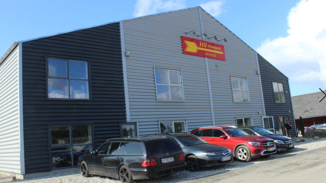 En stor bygning med grå og sort facade, der har vinduer og en rød skilt, samt flere biler parkeret foran.