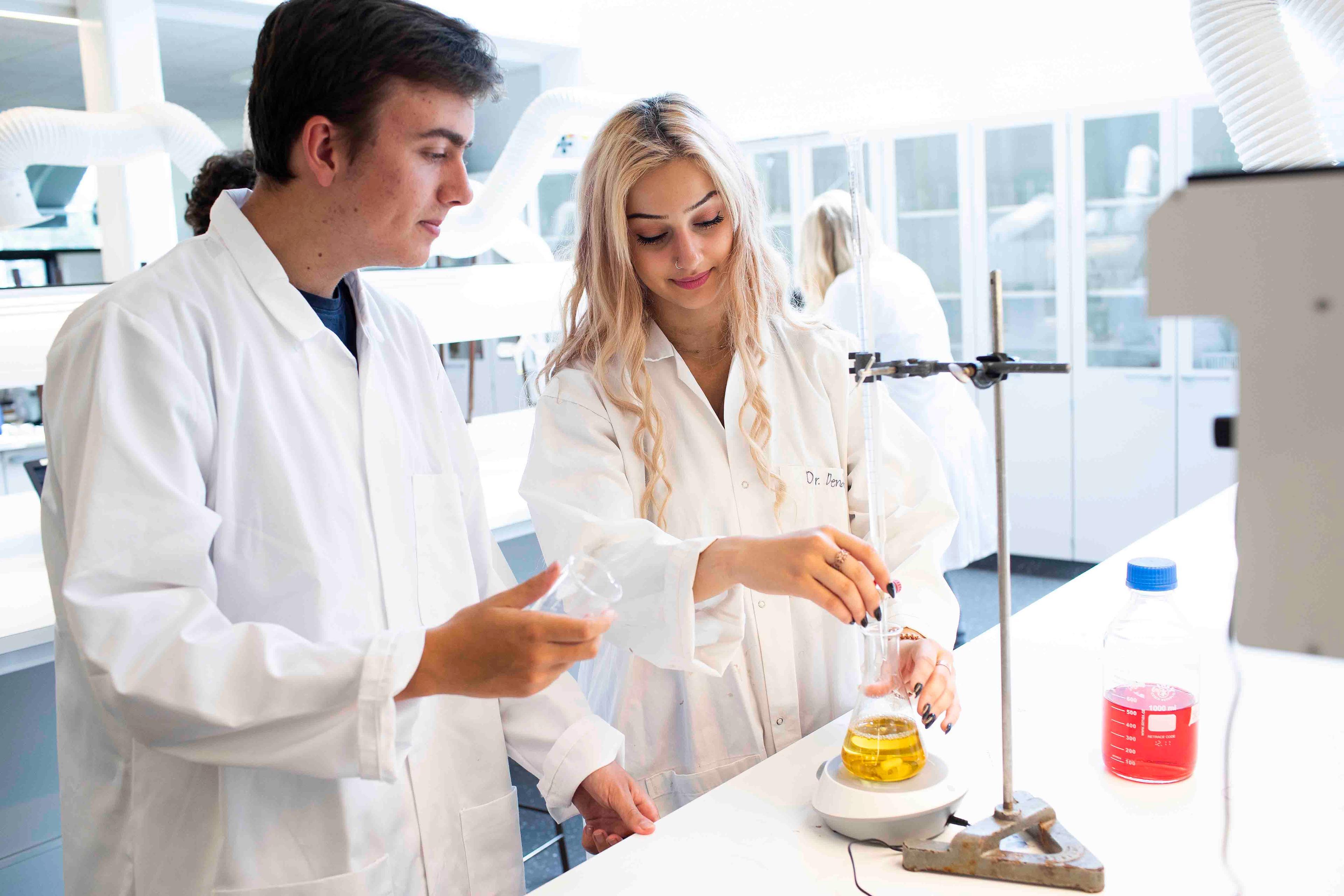 To unge forskere i hvide labcoats, der arbejder med kemiske eksperimenter i et moderne laboratorium.