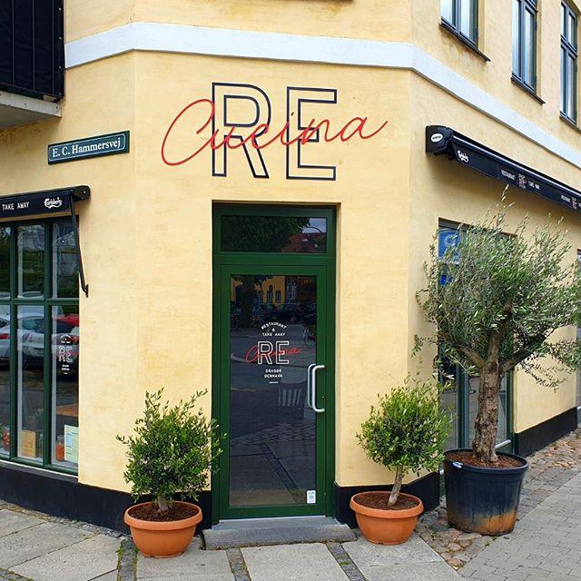 Restaurantens facade med navnet "RE" skrevet i rød skrift på en gul væg, omgivet af planter i potter.