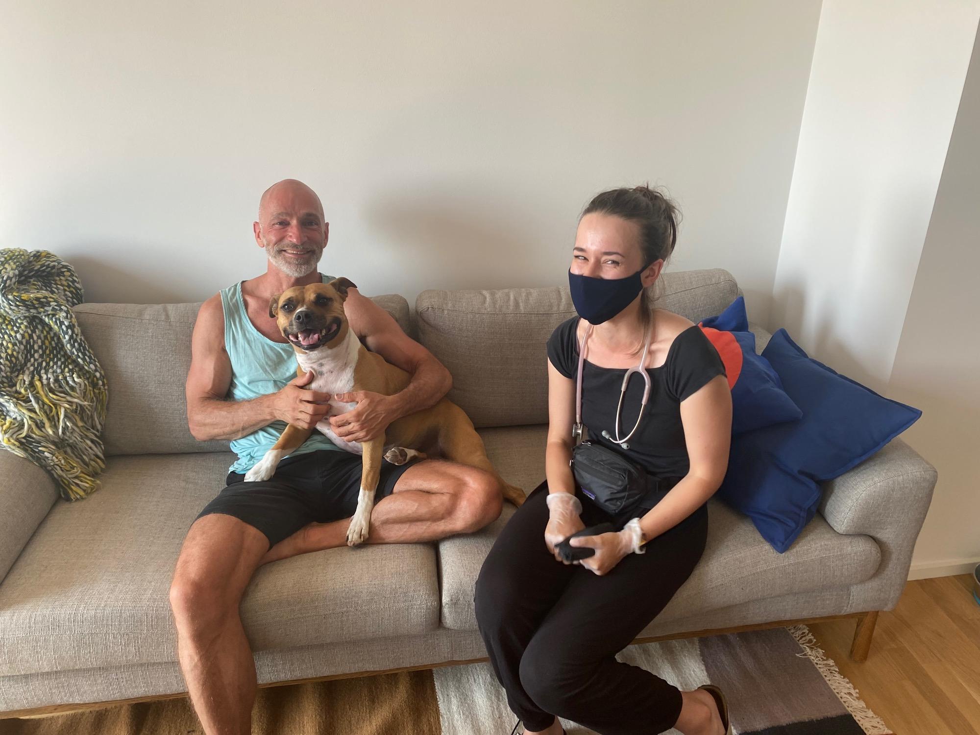 En mand sidder på en sofa med en hund i skødet, mens en kvinde sidder ved siden af med en maske på.