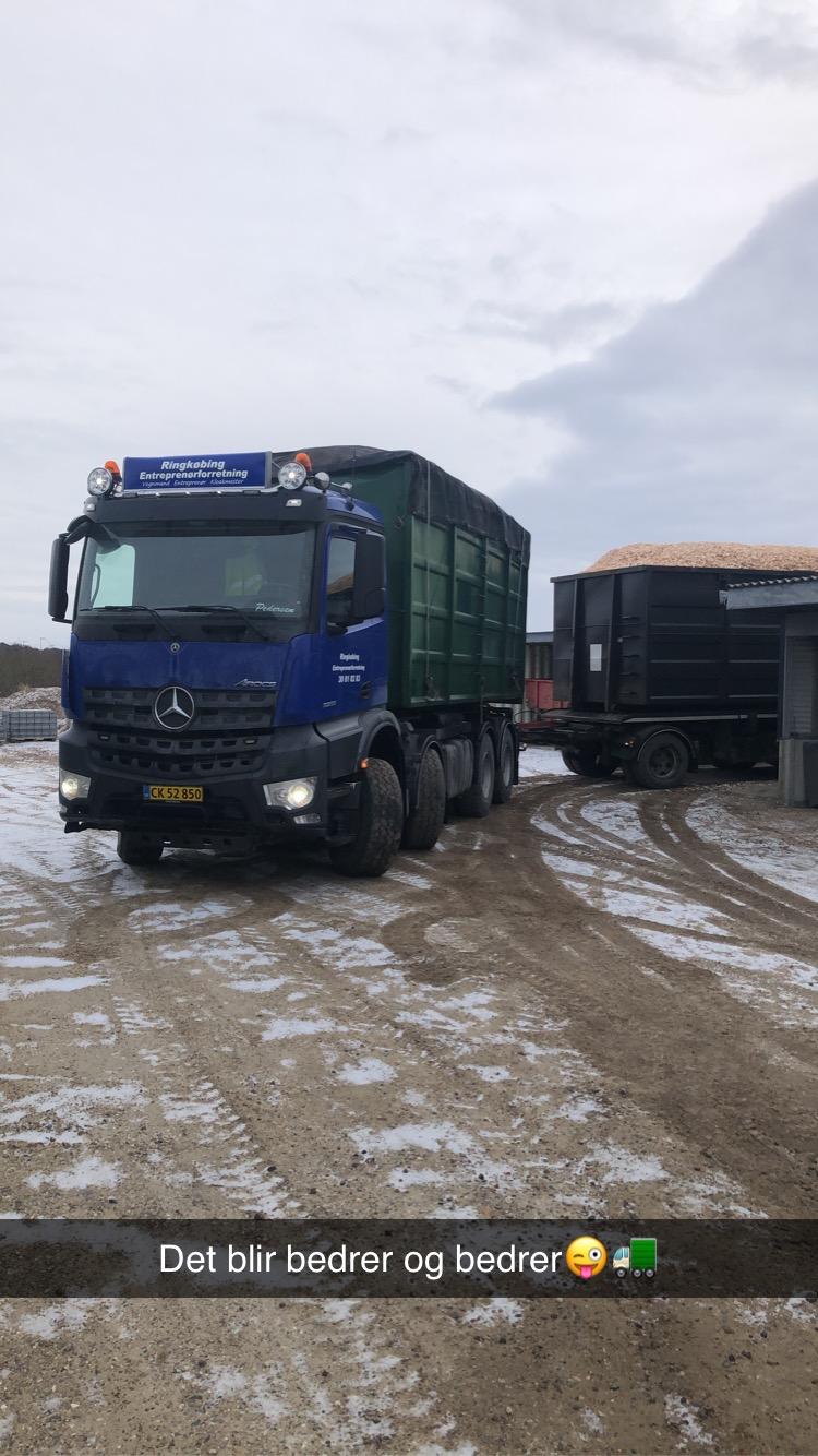 En blå lastbil med en grøn vogn parkeret på en snedækket vej ved en gård.