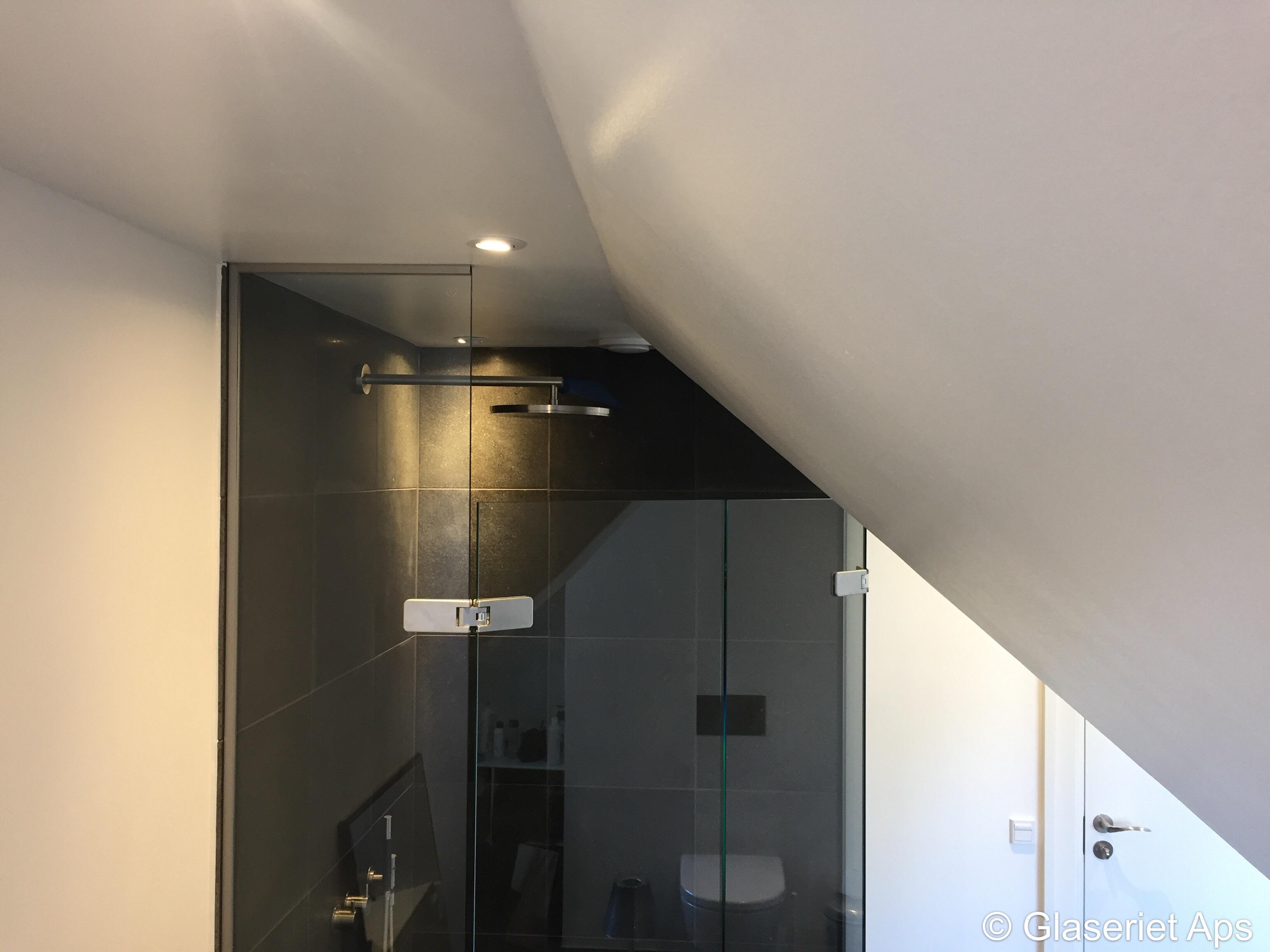 Moderne badeværelse med glasbruser, sorte fliser og skrå loft. Lysarmaturer i loftet.
