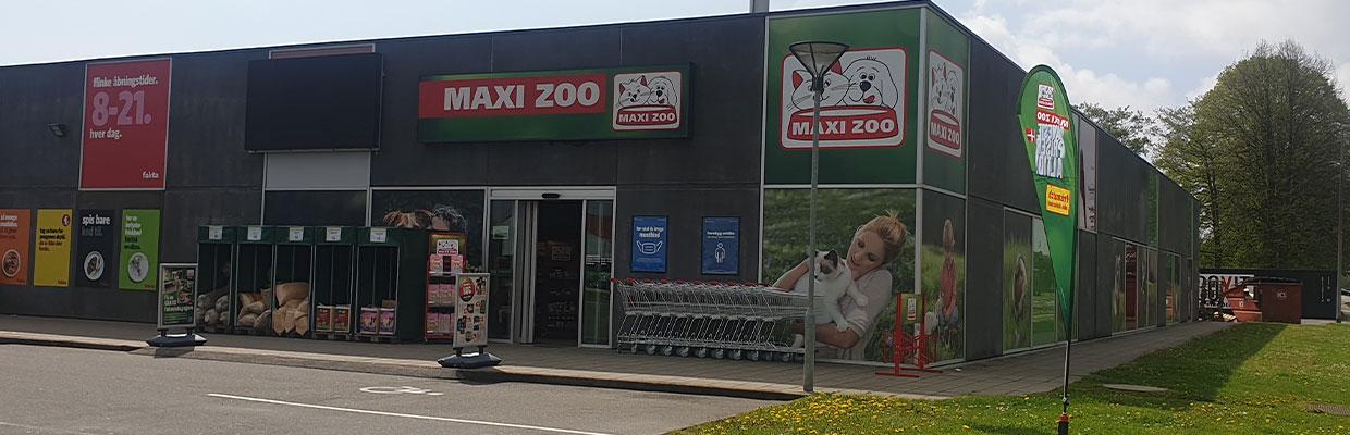 Butikfacade af Maxi Zoo med skilte og indkøbsvogne foran. Solrigt vejr og grønne områder omkring.