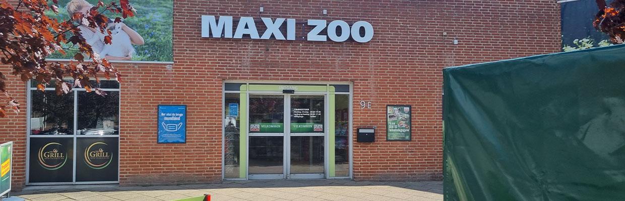 Indgangen til MAXI ZOO med store glasdøre og murstensvæg, omgivet af grønne planter.
