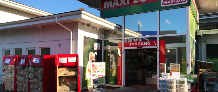 Indgangen til MAXI Zoo med solskin, affaldscontainere og varer synlige udenfor butikken.