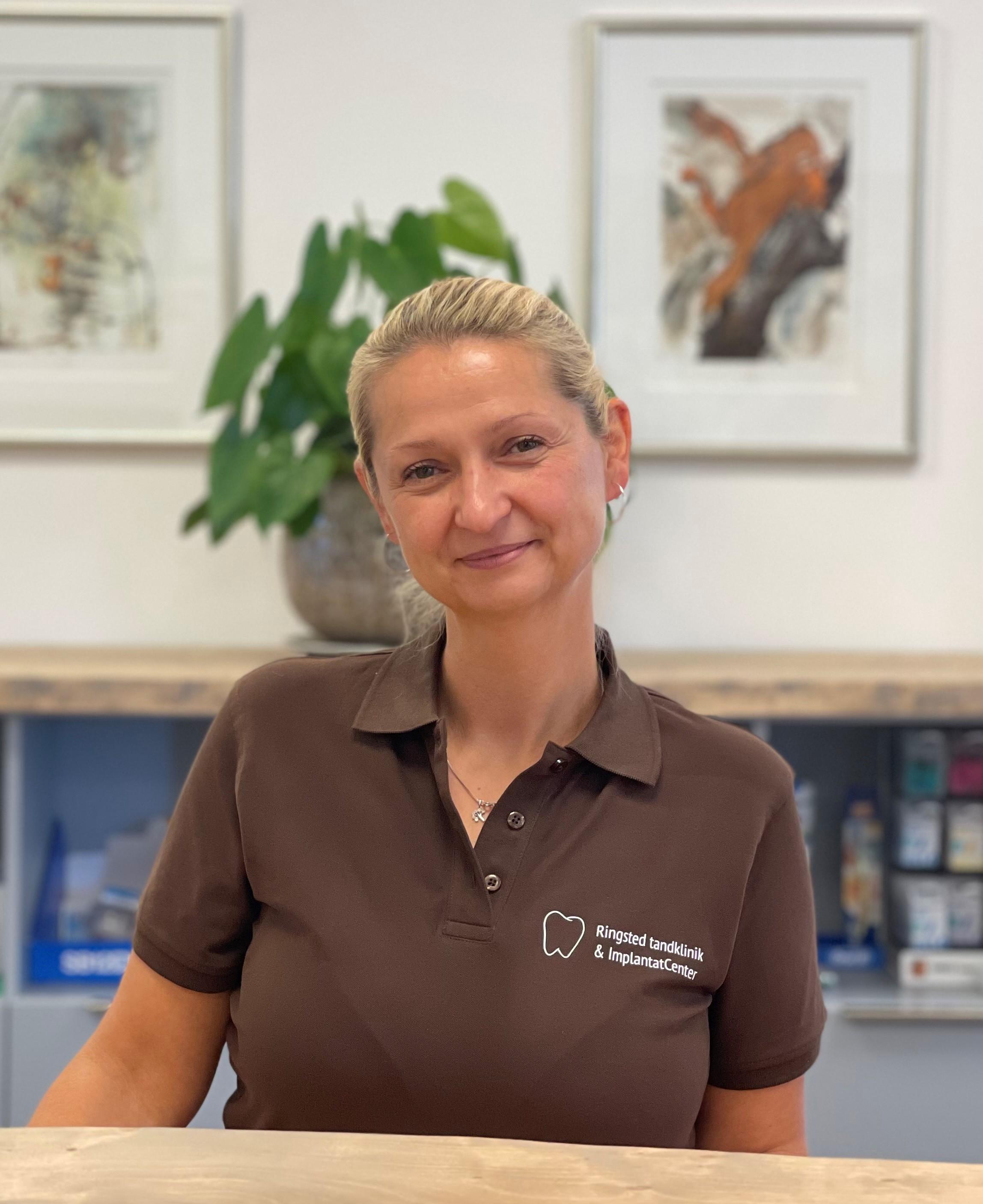 En smilende kvinde i en brun polo t-shirt står ved en reception med planter og kunstværker i baggrunden.
