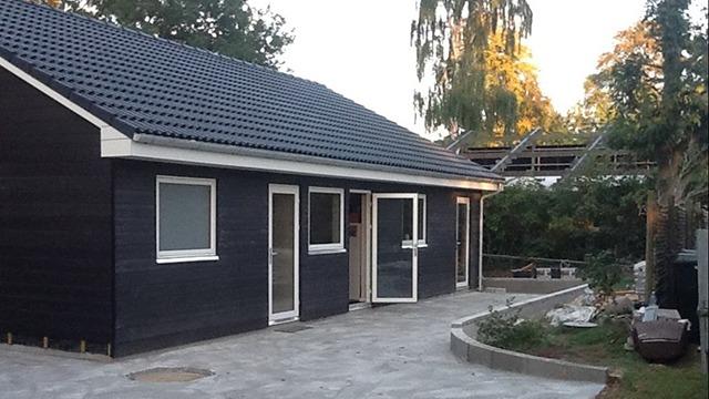 Moderne hus med sort træfacade og flisebelagt indkørsel i en have med træer og buske.