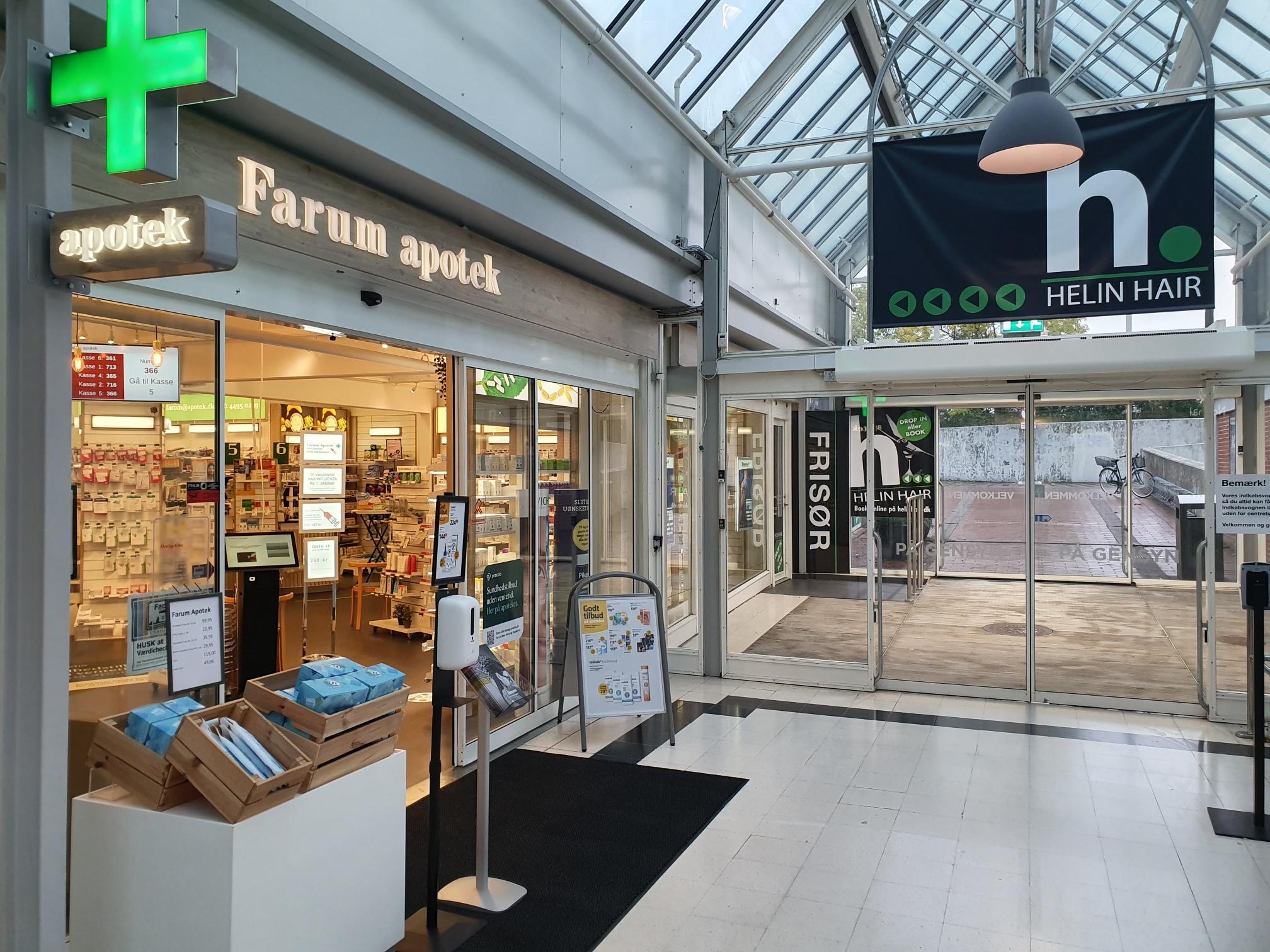 Indgang til Farun apotek med grønne kryds og frisørsalon i baggrunden i et lyst indkøbsområde.