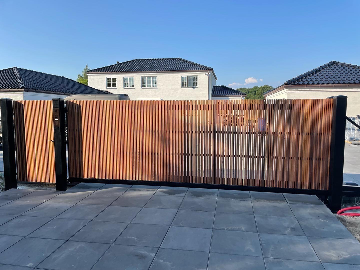 Moderne træport med sorte metalrammer, der står åben foran en grusbelagt indkørsel og huse i baggrunden.
