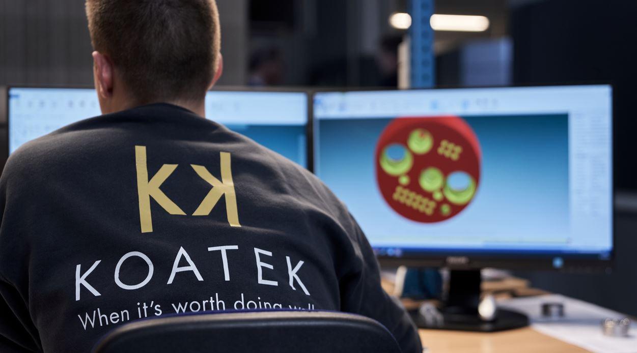 En person i en sweatshirt med teksten "KOATEK" sidder foran to computere, der viser 3D-modeller.