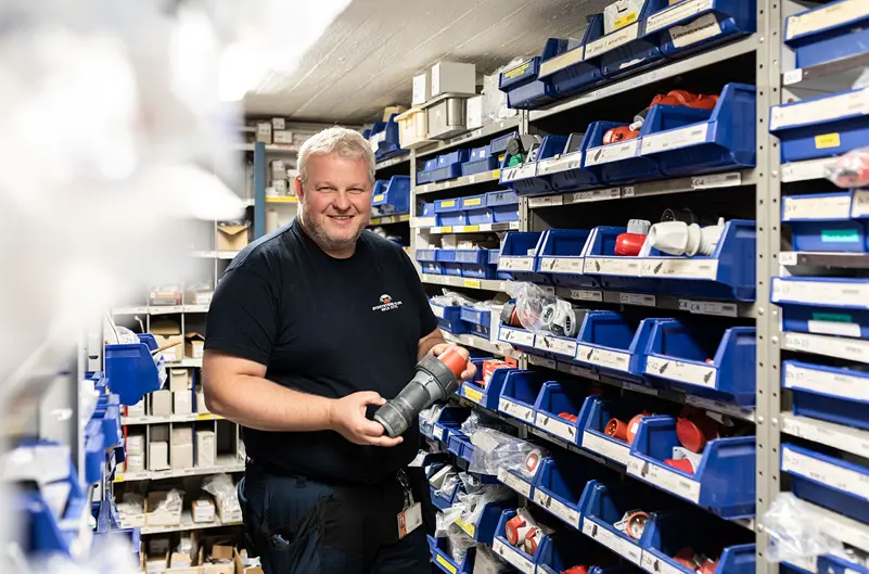 En smilende mand holder en rørdel i et lager fyldt med blå opbevaringskasser og værktøj.
