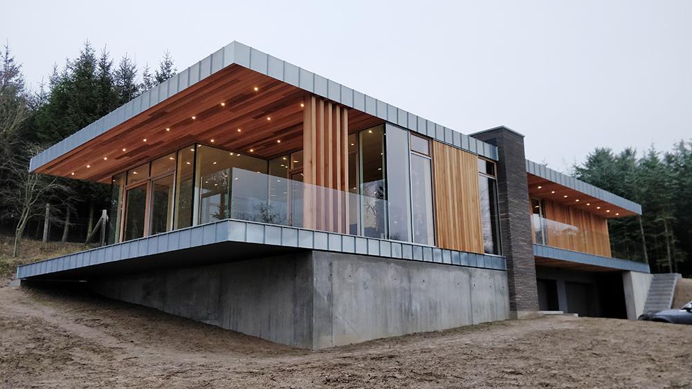 Moderne hus med træfacade og glasvægge, beliggende i skovområde, med skrånende grund og udendørs terrasse.