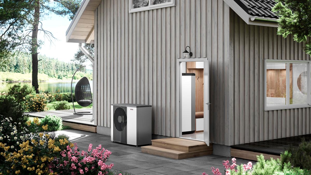 Et moderne hus med grå træbeklædning, en terrasse, blomsterbed og en varmepumpe ved siden af indgangen.