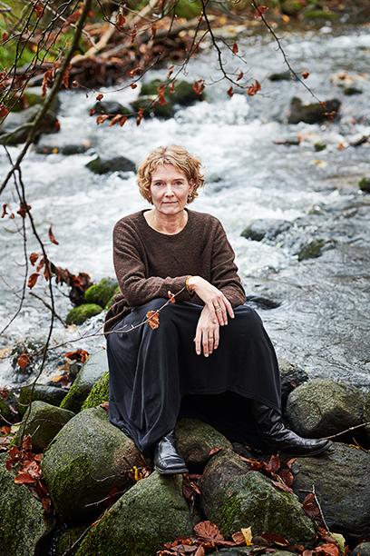 Kvinde sidder på sten ved en strøm med vand, iført en brun trøje og sort nederdel, omgivet af efterårsblade.