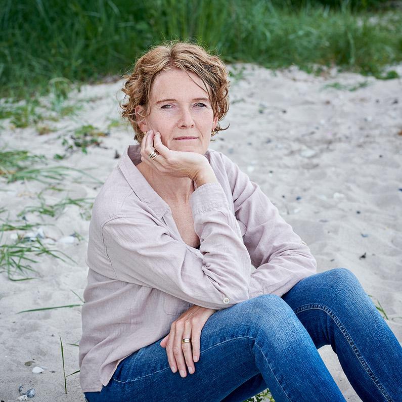 Kvinde sidder på stranden med hånden under hagen, iført en lyserød skjorte og jeans, omgivet af græs og sand.