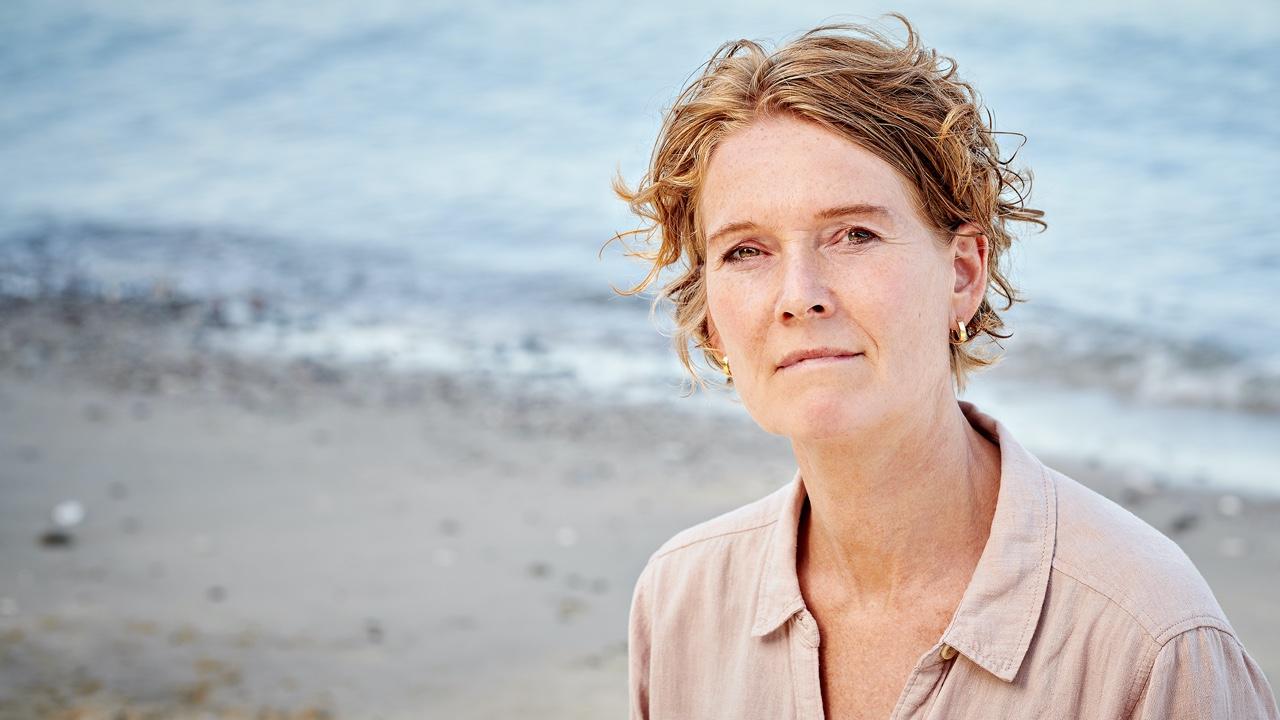 Portræt af en kvinde med kort, krøllet hår, der sidder ved stranden med havet i baggrunden.