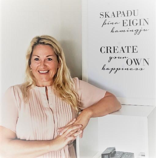 En smilende kvinde med langt, lyst hår står ved en væg med inspirerende citater om lykke.