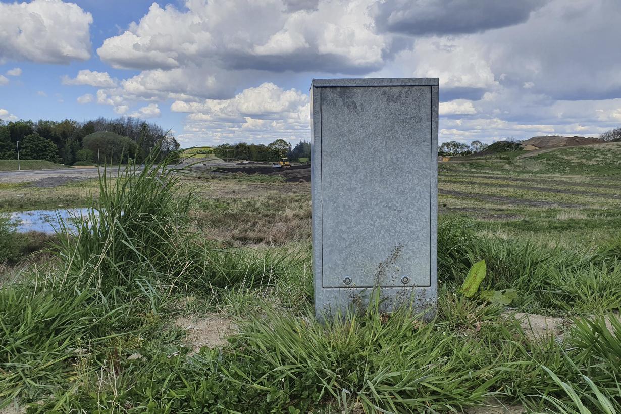 En grå stenmonument står i en grøn eng med skyer i baggrunden og marker i horisonten.