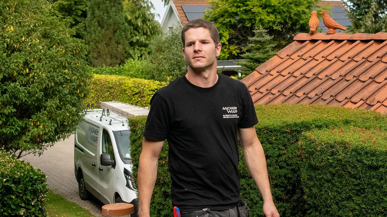 En mand i sort t-shirt går på en sti mellem buske, med en hvid varevogn i baggrunden og et hus med rødt tag.