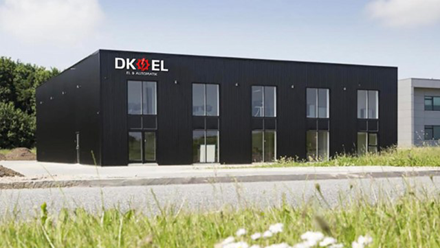 Moderne sort bygning med DK EL-logo, beliggende ved en vej omgivet af græs og træer.