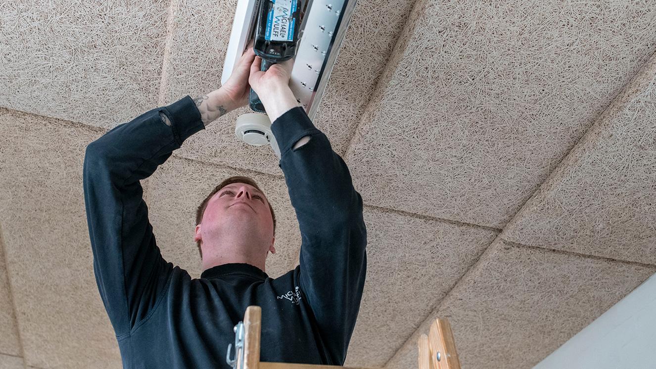 En håndværker står på en stige og installerer en loftventilator i et rum med akustiske lofter.