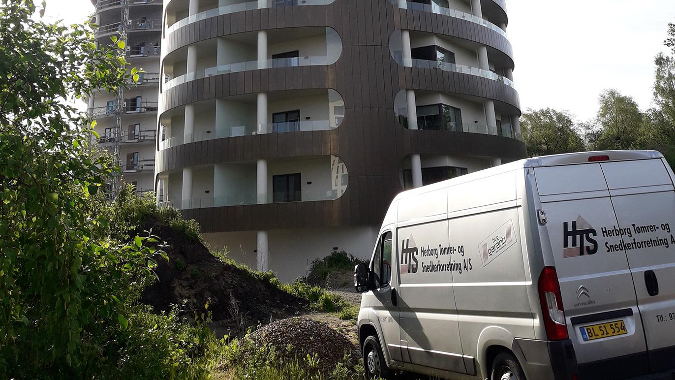 En hvid varevogn parkeret foran en moderne bygning med flere etager og en grøn beplantning omkring.