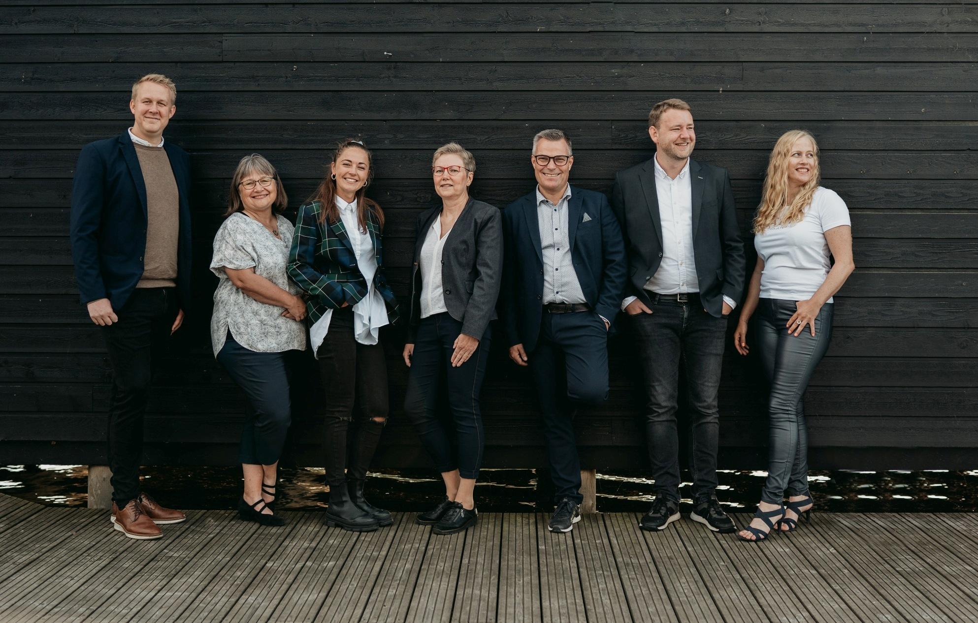 Syv personer står i en række foran en sort væg, iført forskellige professionelle outfits og smilende.