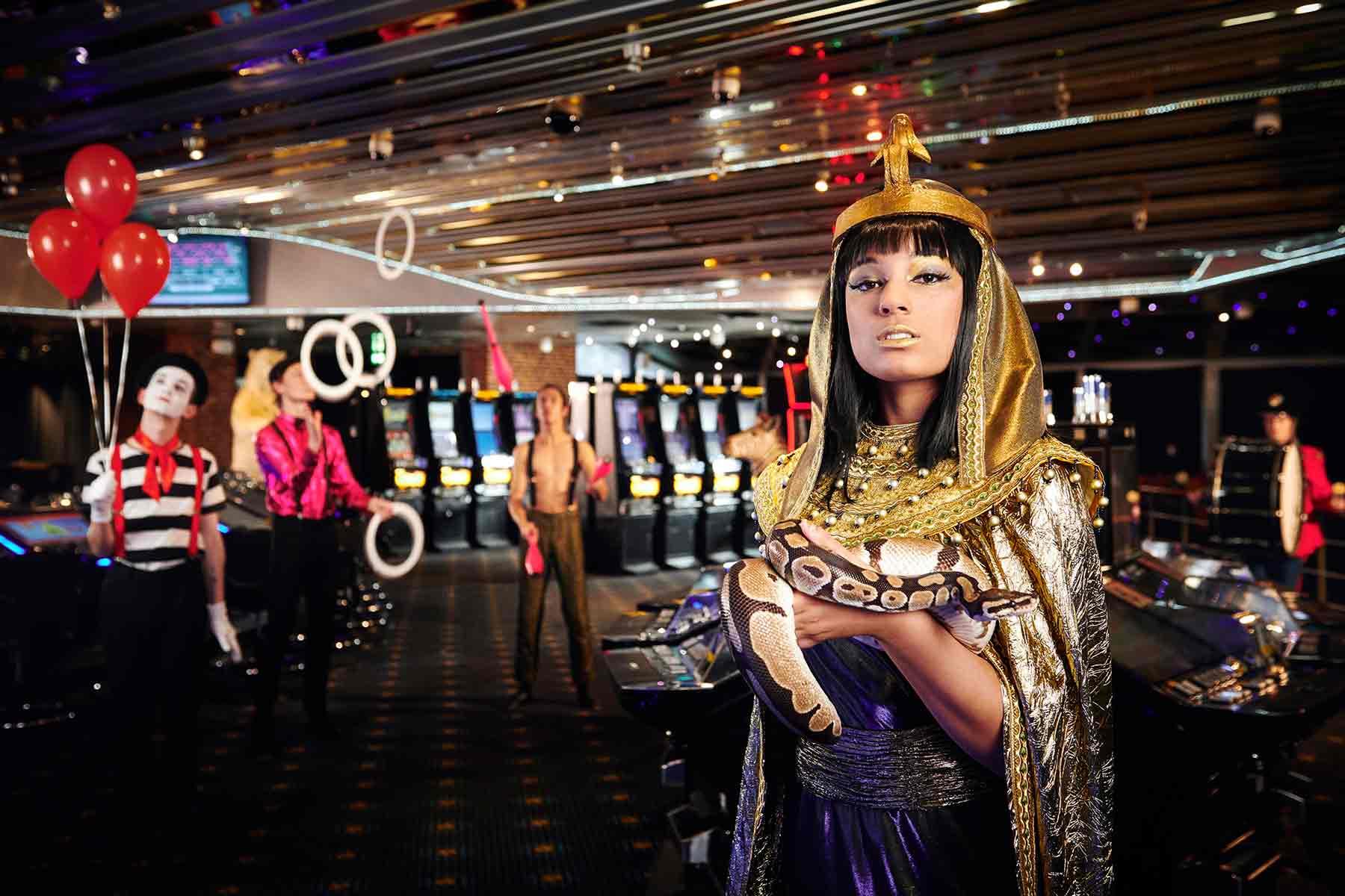 En kvinde klædt som Cleopatra står i et kasino med underholdning og spillemaskiner i baggrunden.