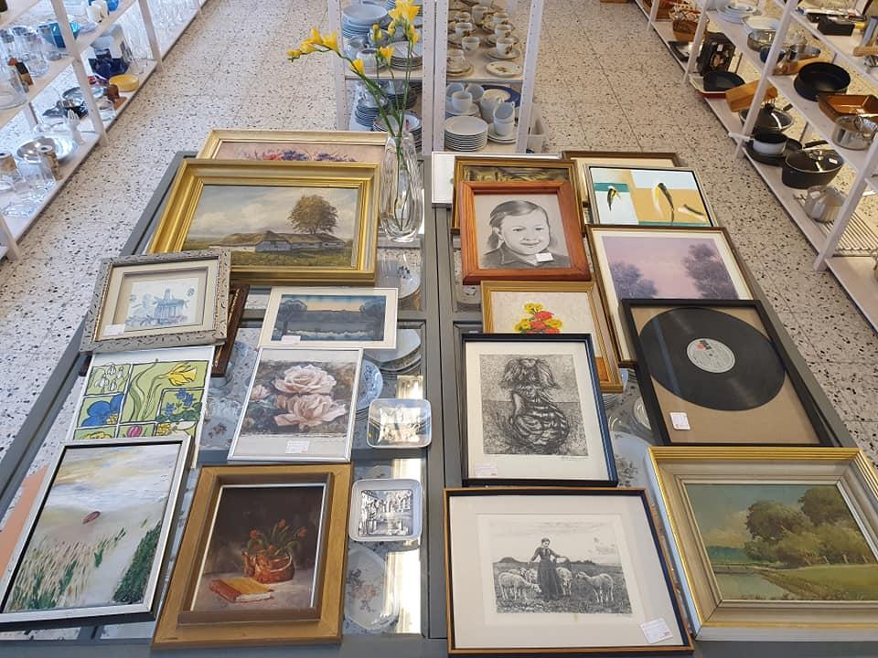 Bord med forskellige rammer og billeder, herunder malerier, fotografier og en plade, i en butik.