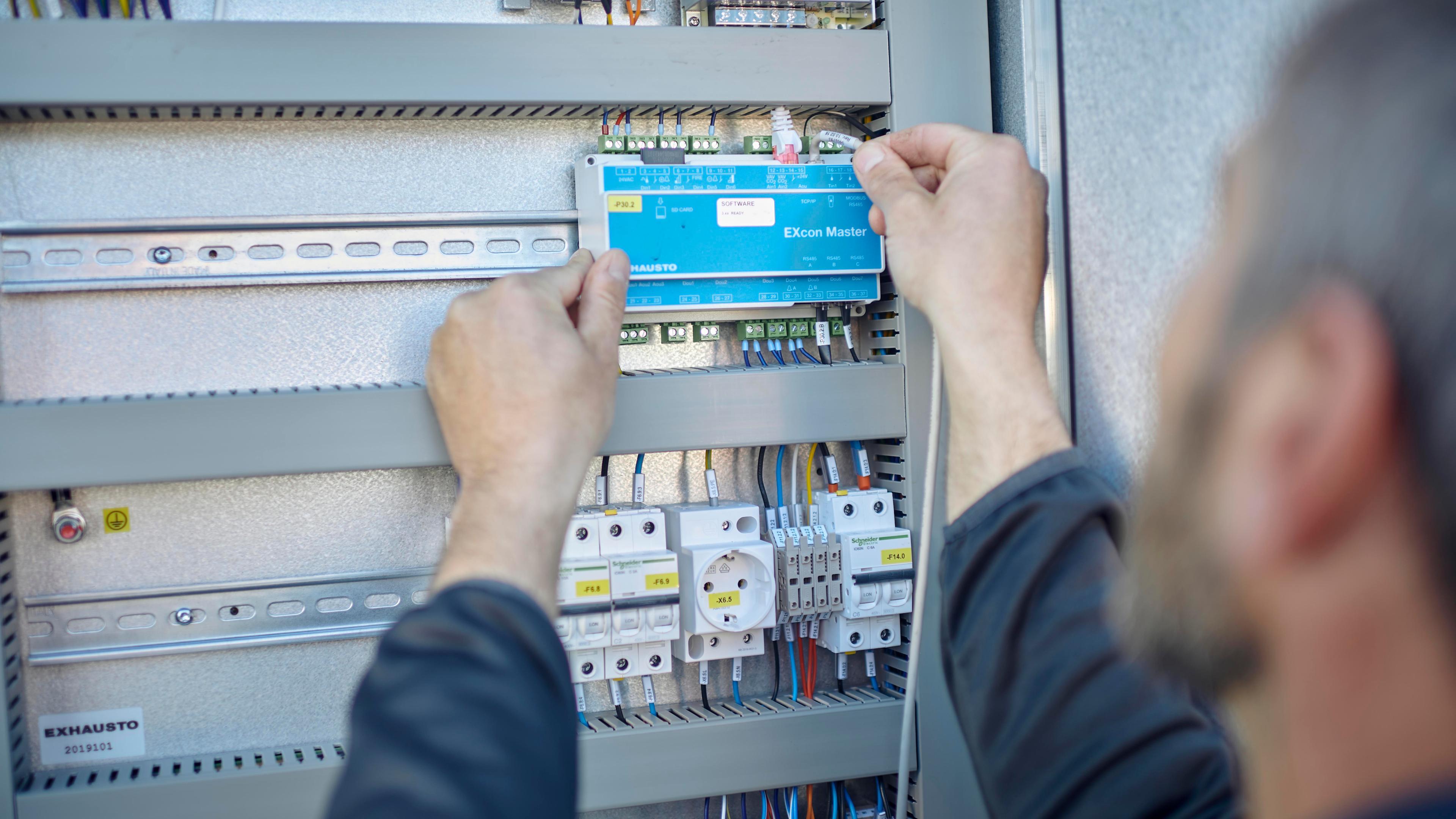 En person installerer en elektrisk komponent i et skab med ledninger og sikringer.
