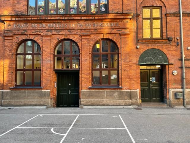 En rød murstensbygning med store vinduer og en gård med en basketballbane foran.