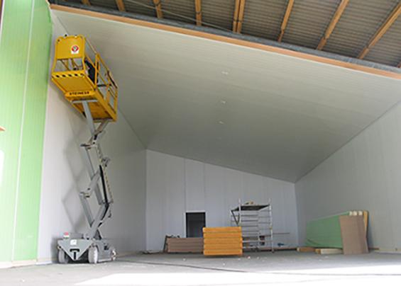 En gult liftarbejde i et stort, tomt rum med hvide vægge og en skrå loft. Byggematerialer er synlige på gulvet.