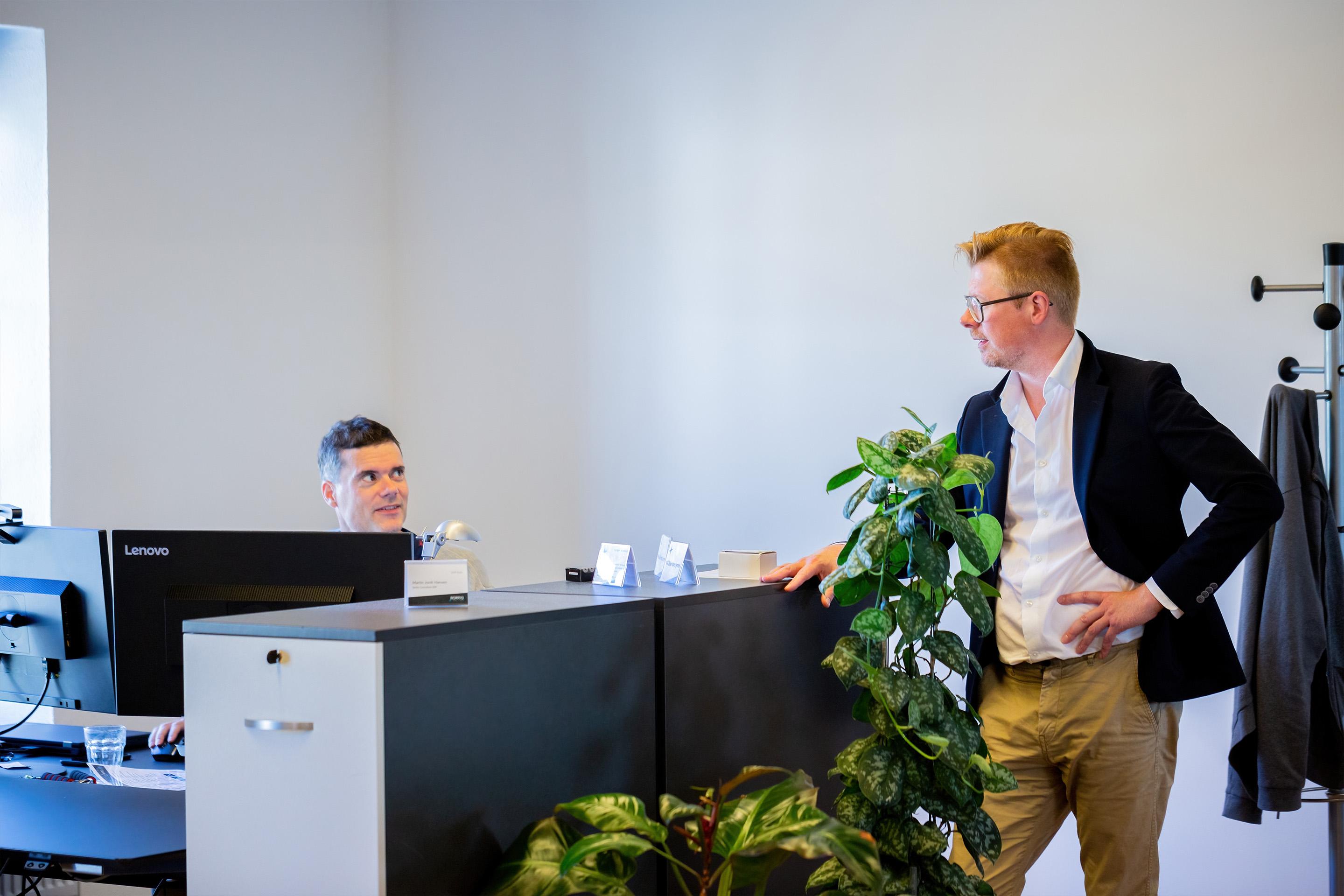 To mænd i et kontor, hvor den ene står ved en reception og den anden sidder ved en computer. Grøn plante i forgrunden.
