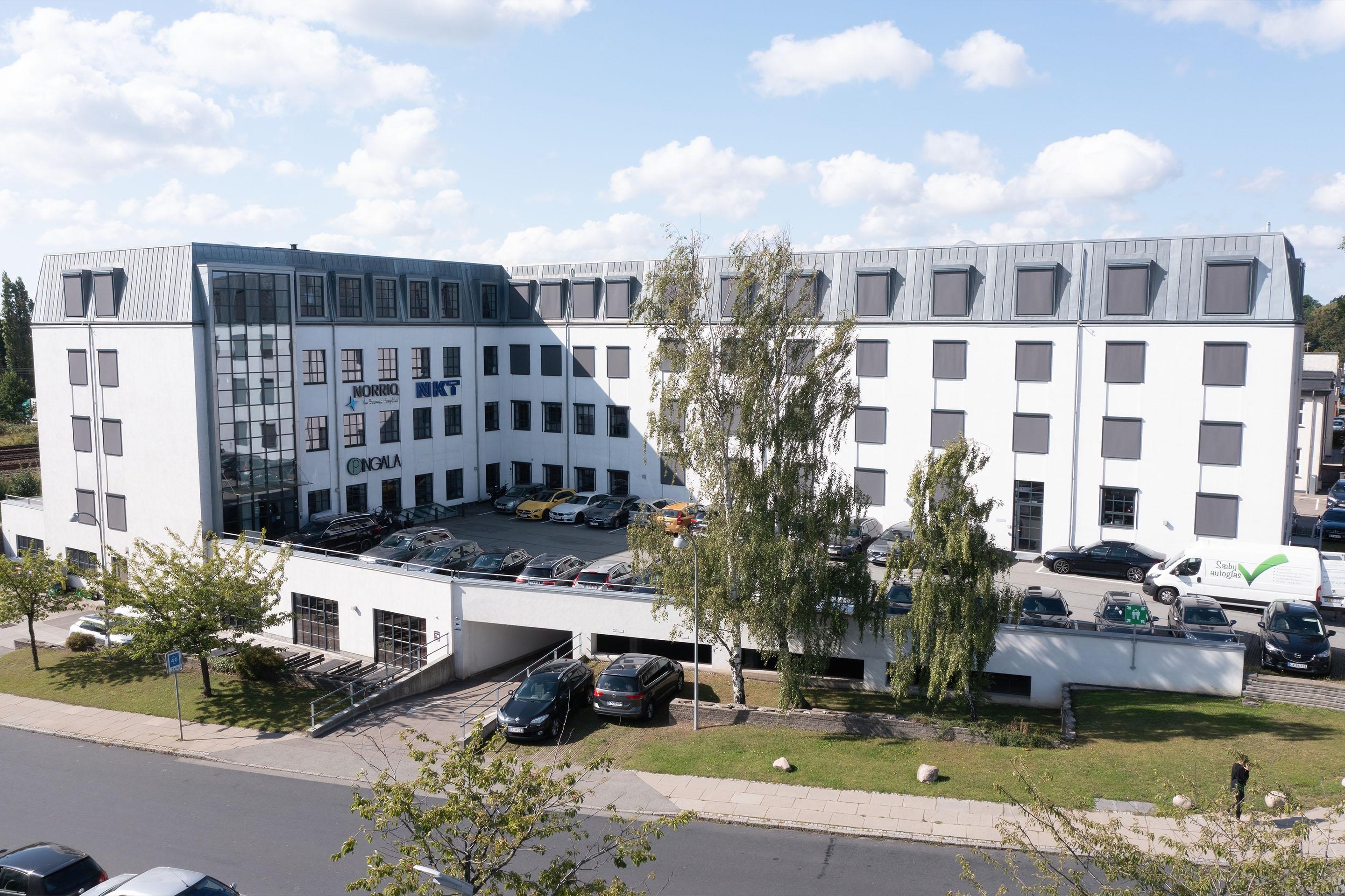 Moderne kontorbygning med flere etager, parkeringsplads og grønne områder omkring.