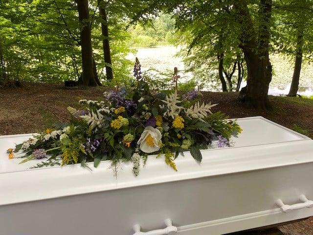 En hvid kiste dekoreret med farverige blomsterarrangementer i en skov med grønne træer.