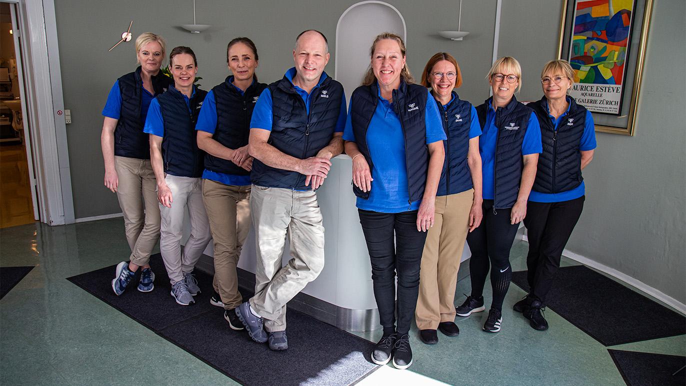 Et team af ni personer i blå skjorter og sorte veste står sammen i en moderne reception.