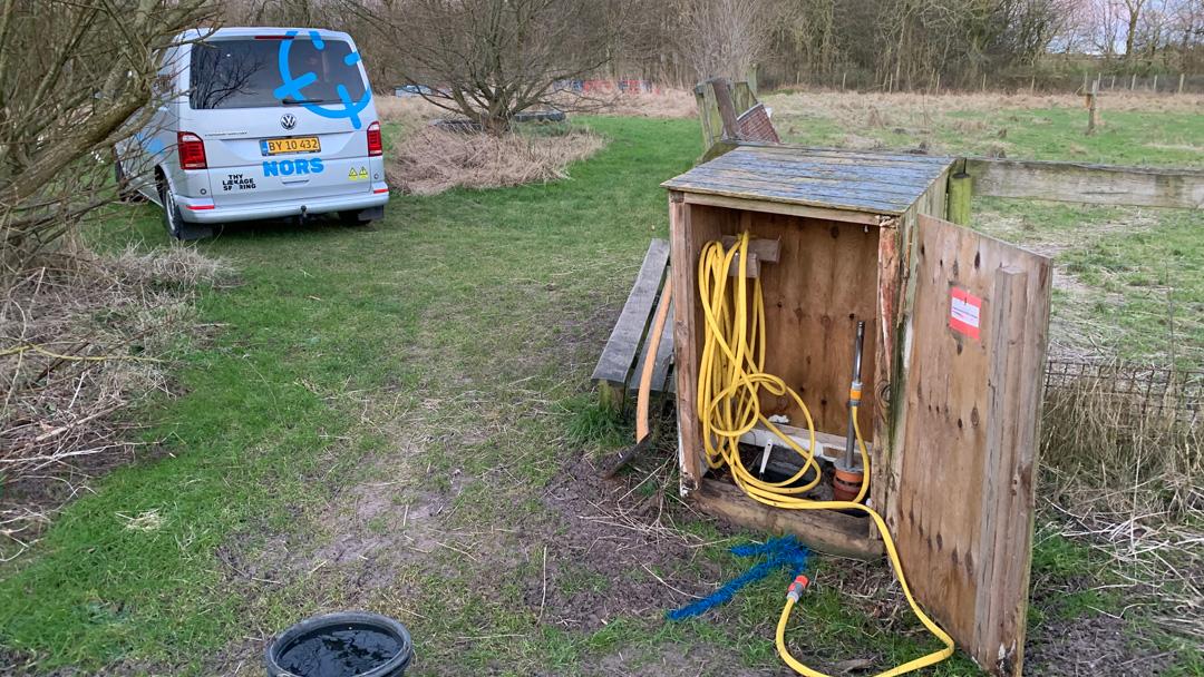 En hvid varevogn parkeret ved en træhytte med slanger og udstyr i en græsset park.