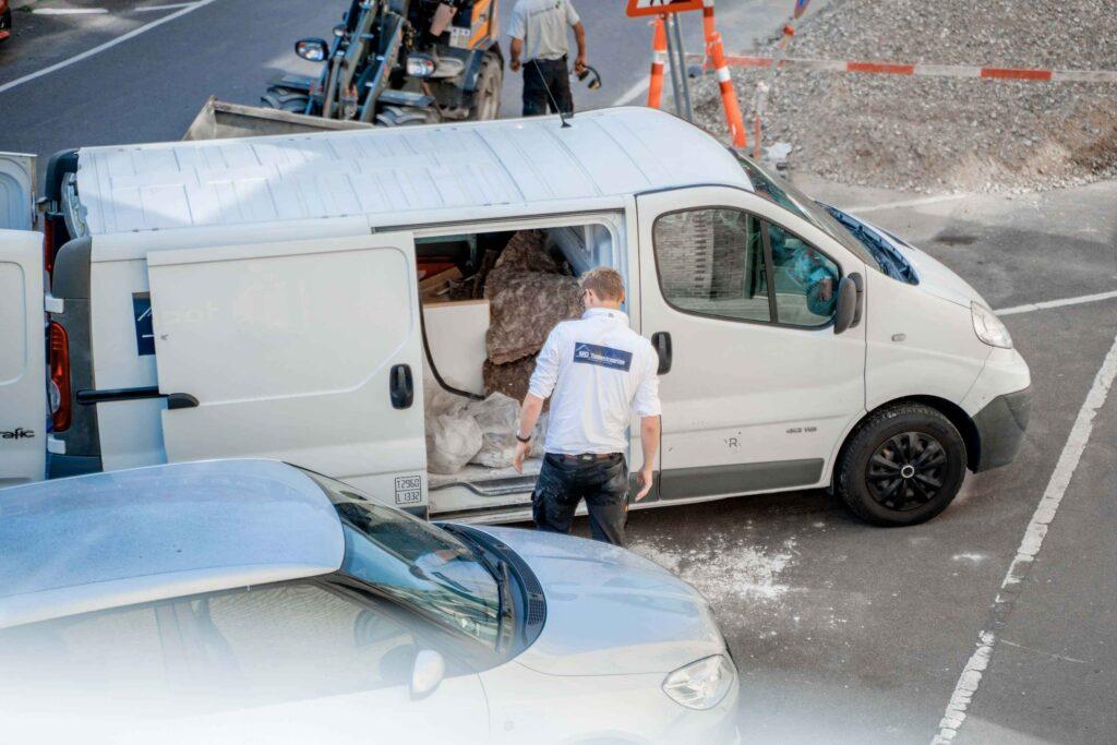 En mand står ved en hvid varevogn, der er fyldt med materialer, mens biler parkerer i nærheden.