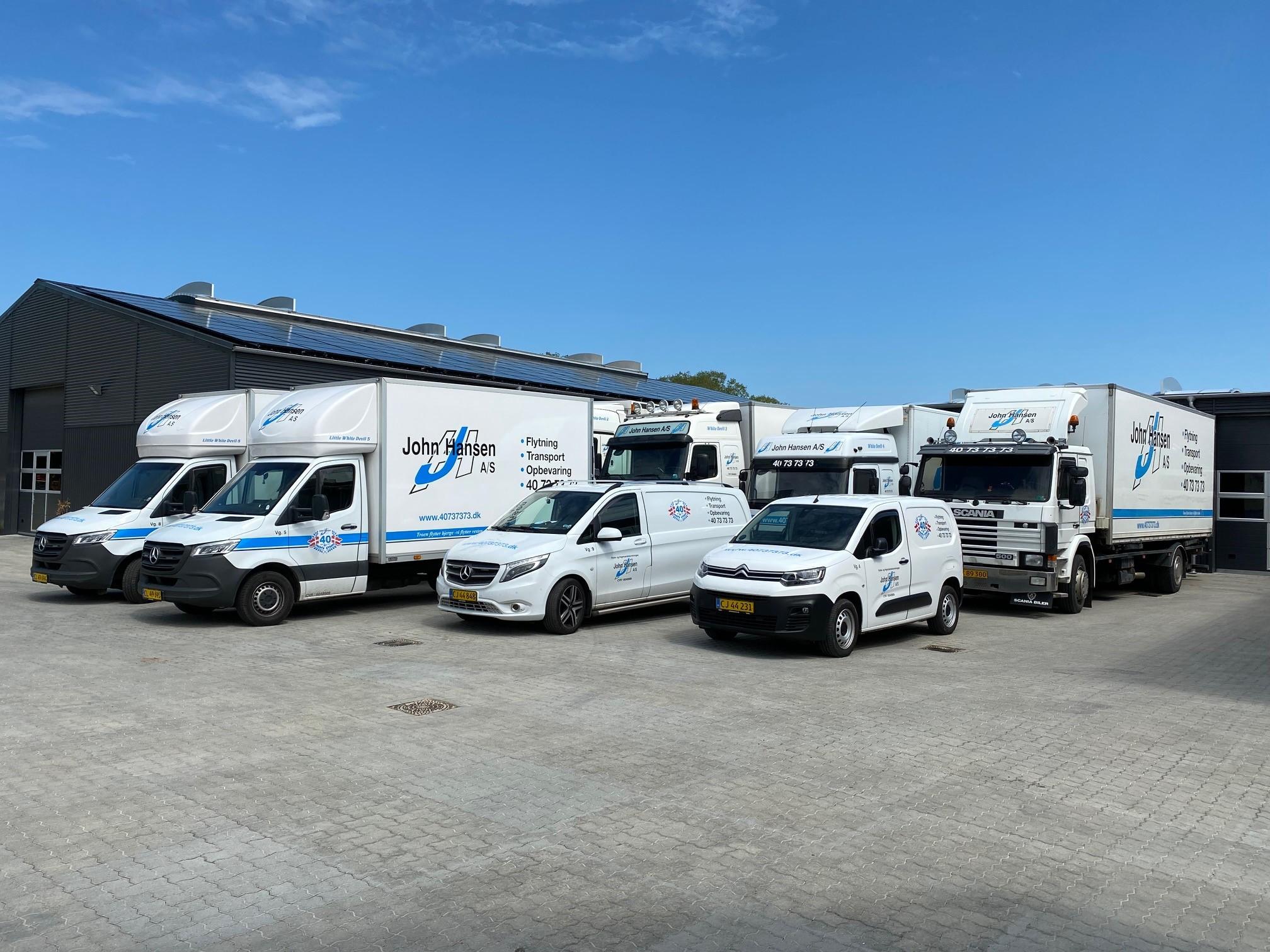 Flere lastbiler og varevogne parkeret foran en bygning på en solrig dag.