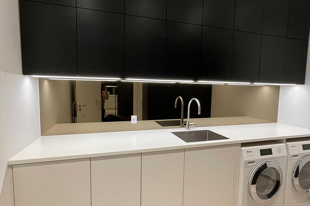 Moderne vaskerum med sort skab, hvid bordplade, vask og vaskemaskiner. Spejlrefleksioner i baggrunden.