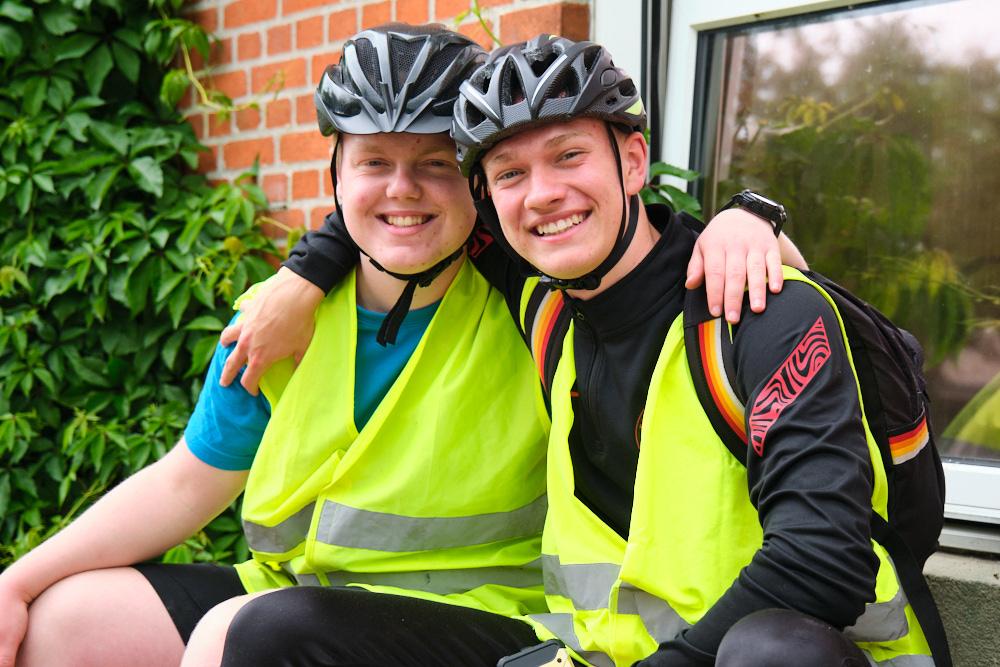 To unge cyklister iført hjelme og refleksveste, der smiler og sidder sammen foran en bygning.