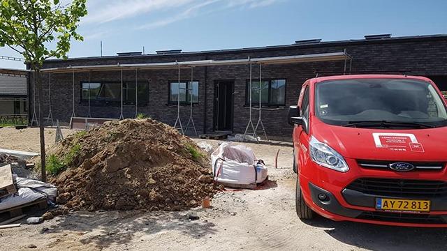 Byggeri af et hus med en rød varevogn og en bunke jord foran. Scenen viser stillads og byggeaktiviteter.