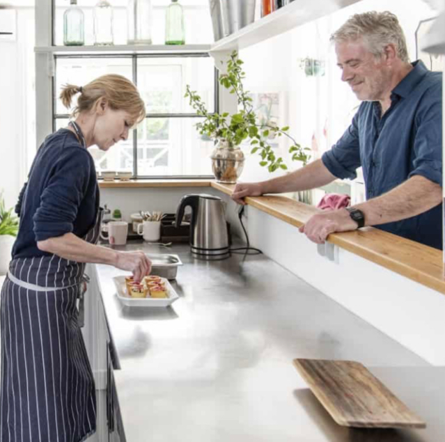 En kvinde i stribet forklæde forbereder mad på en moderne køkkenbænk, mens en mand smilende ser på.