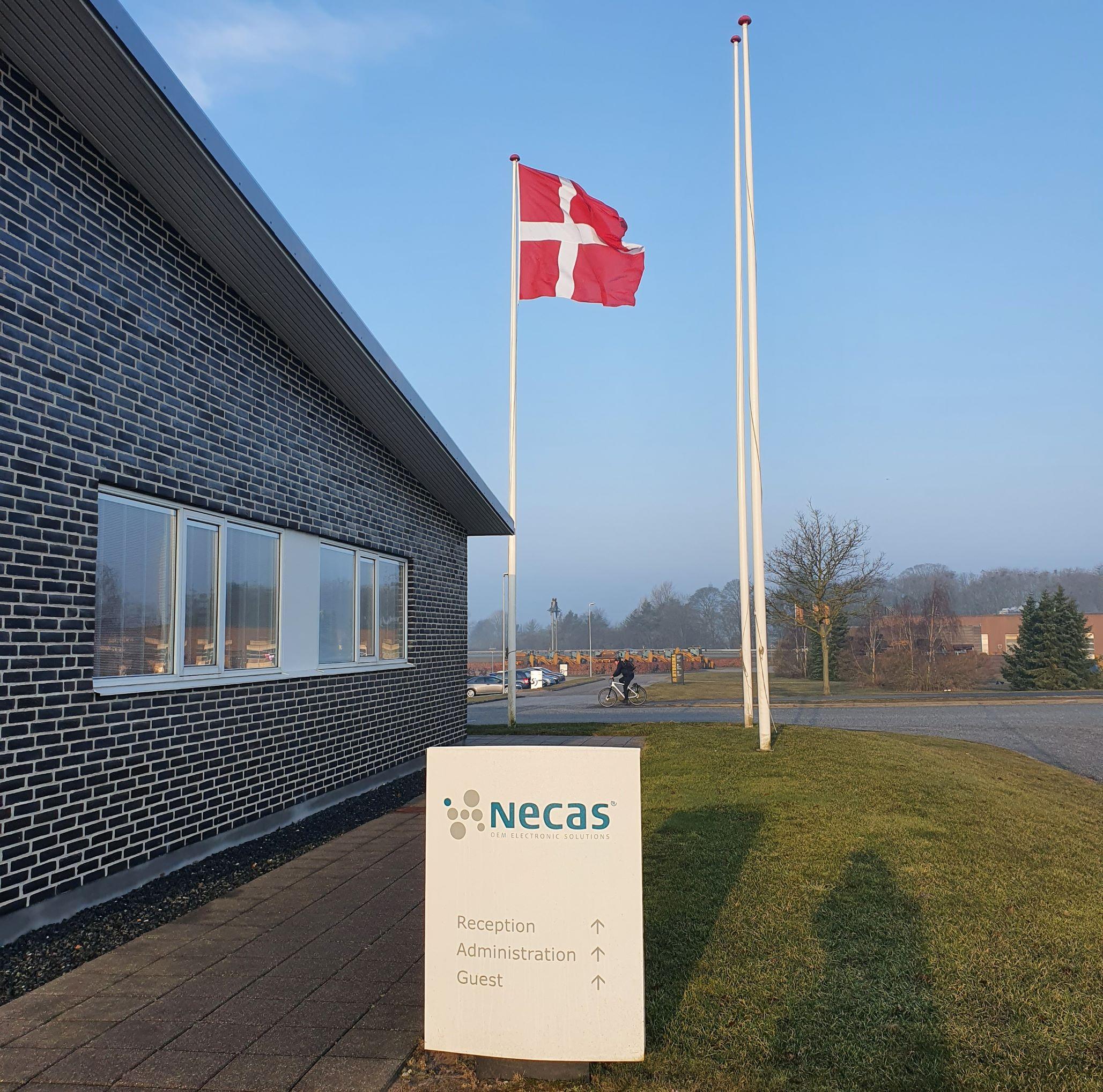 Bygning med det danske flag og et skilt for NECAS i morgenlys.