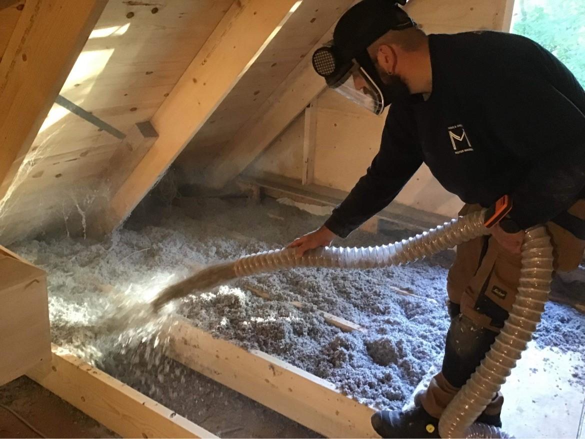 En håndværker, der blæser isoleringsmateriale ind i et loft med en slange, omgivet af støv og træbjælker.