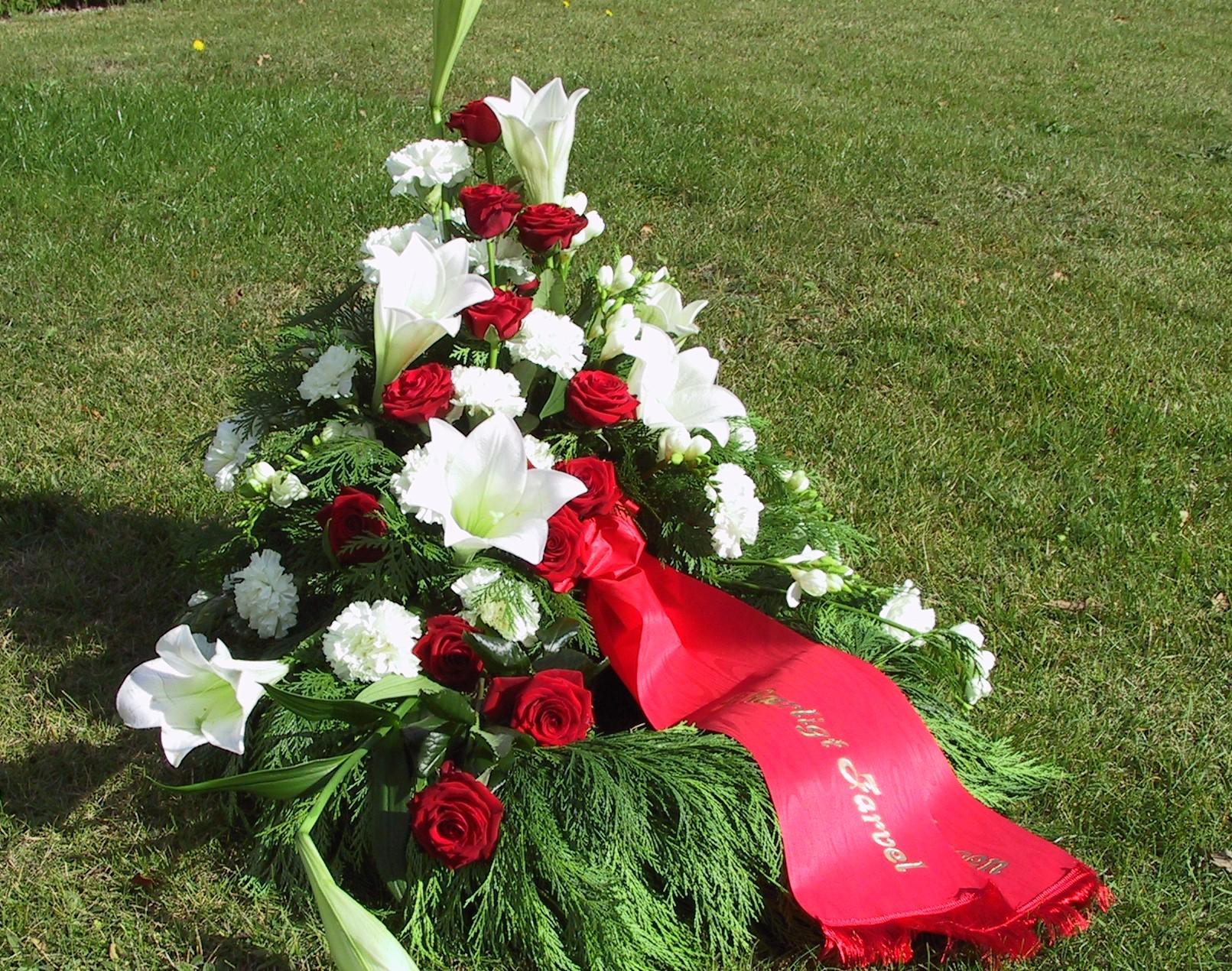 En smuk blomsterkrans med hvide liljer, røde roser og grønne blade, dekoreret med et rødt bånd.
