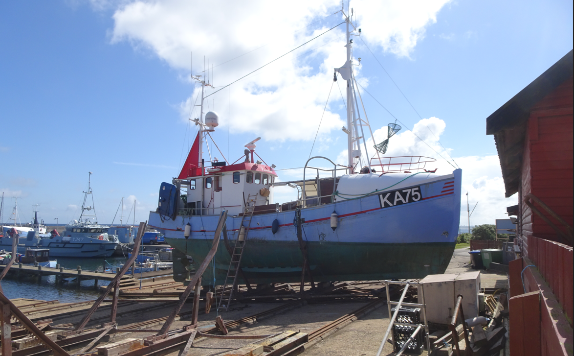 Fiskebåd KA75 på land ved havnen, omgivet af træplanker og bygninger. Blå himmel med skyer i baggrunden.