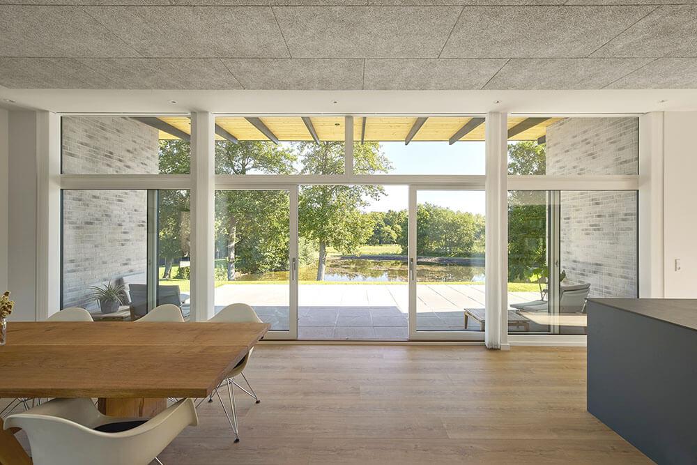 Moderne stue med store vinduer, der giver udsigt til en have og terrasse ved vandet. Træbord og minimalistisk indretning.