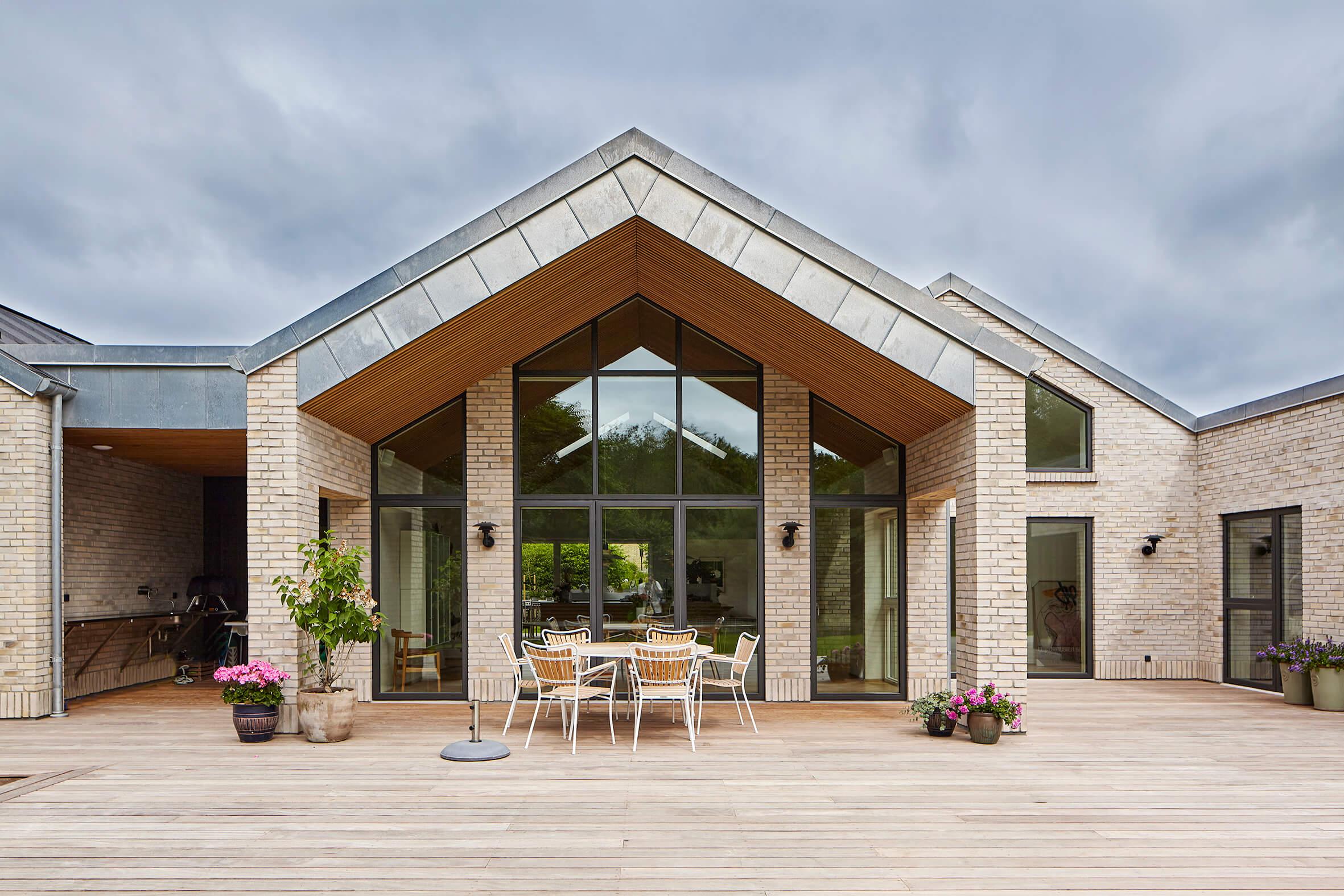 Moderne hus med skråtag, store vinduer og en træterrasse med spiseplads og blomsterkrukker.