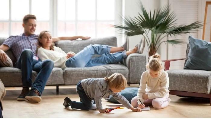 Familie i stuen, hvor børn leger på gulvet, mens forældre slapper af på sofaen. Grøn plante i baggrunden.